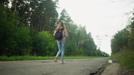 young woman with backpack walking alone on long rural tarred road lined with lush green trees, peaceful atmosphere, forest surroundings, casual outfit with hoodie and jeans, overcast sky above