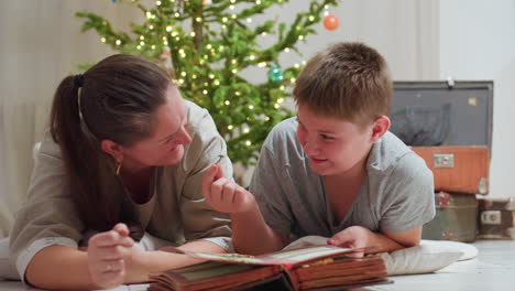 woman and son lying on marble floor smiling joyfully while picking little stick from open vintage book cozy festive home environment background with christmas tree playful moment together