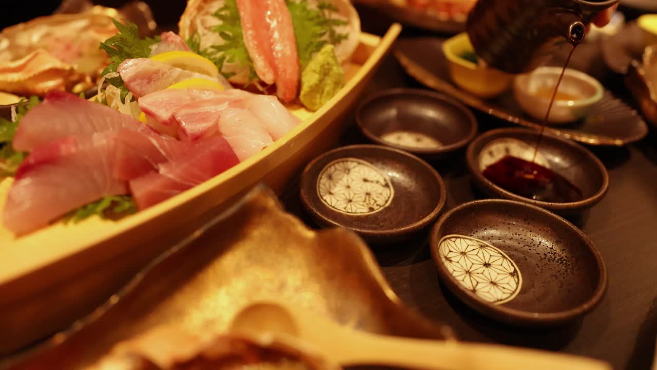 Una mano versa salsa di soia scura in ciotole di ceramica accanto a un vassoio di legno a forma di barca per sashimi.