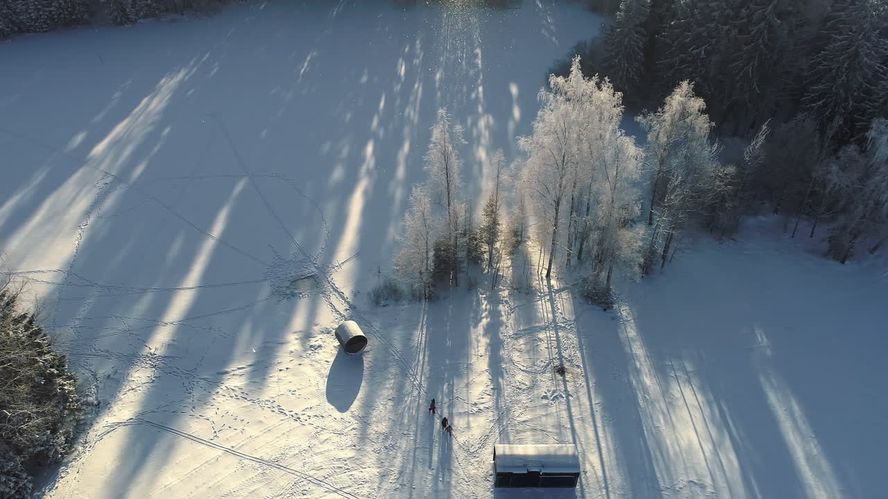 toma aérea de arriba hacia abajo del paisaje nevado de invierno con cabaña de madera iluminada por la luz del sol en la mañana - grupo de personas caminando en un campo rural nevado