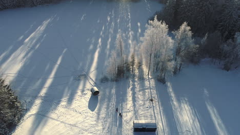 Toma-Aérea-De-Arriba-Hacia-Abajo-Del-Paisaje-Nevado-De-Invierno-Con-Cabaña-De-Madera-Iluminada-Por-La-Luz-Del-Sol-En-La-Mañana---Grupo-De-Personas-Caminando-En-Un-Campo-Rural-Nevado