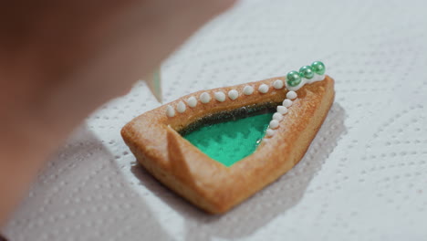 close-up of cookie decoration in progress with white icing and green beads, cookie with intricate piping details placed on white tissue paper, showcasing meticulous baking