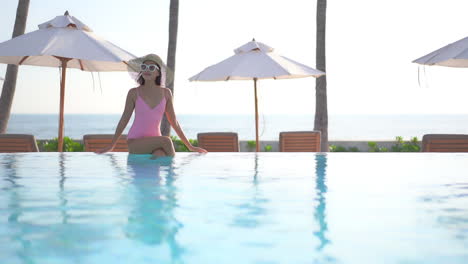 Una-Mujer-Joven-Y-Guapa-Con-Un-Enorme-Sombrero-Para-El-Sol-Y-Gafas-De-Sol-Se-Sienta-Al-Borde-De-La-Piscina-De-Un-Complejo-Con-Los-Pies-En-El-Agua