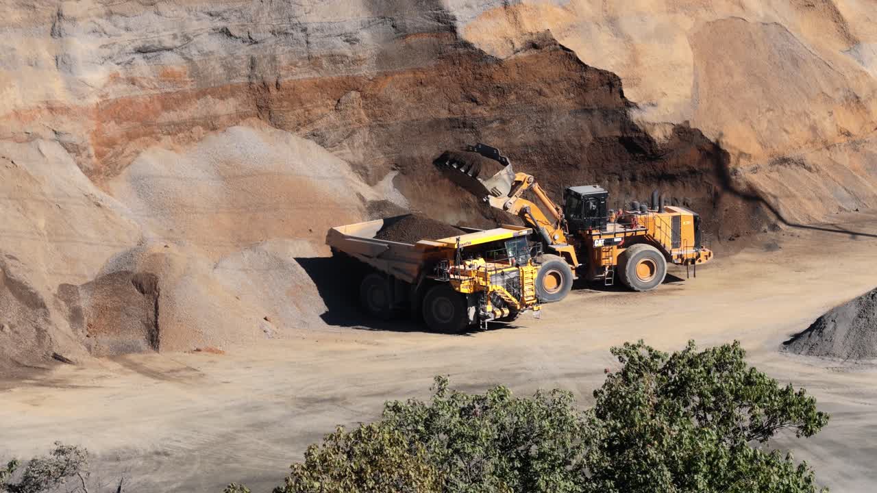 Dump truck parked beside wheel loader as fill and aggregate is loaded for transport, aerial pullback