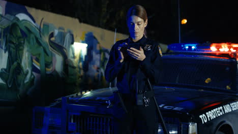 policewoman standing at night on street at police car and tapping on phone