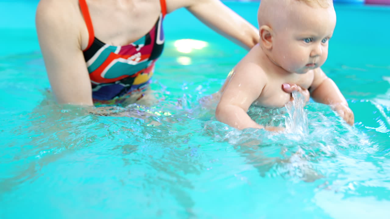 Happy Caucasian baby boy is learning to swim. Child is held by the woman moving him in water.