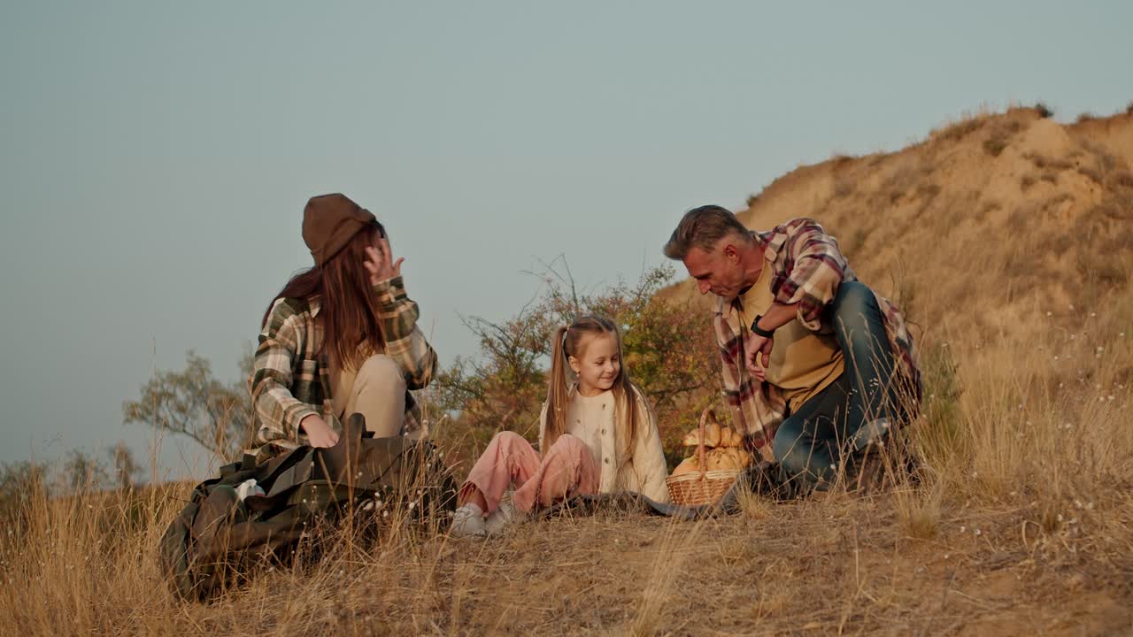 Happy brunette man with gray hair in a brown plaid shirt along with his wife and a brunette woman in a green plaid shirt and little daughter are relaxing on their picnic outside the city in the summer