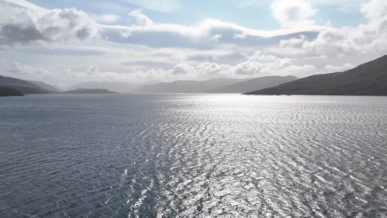 Seabirds fly over sunlit loch, lighthouse, and dramatic clouds in the Scottish Highlands panorama