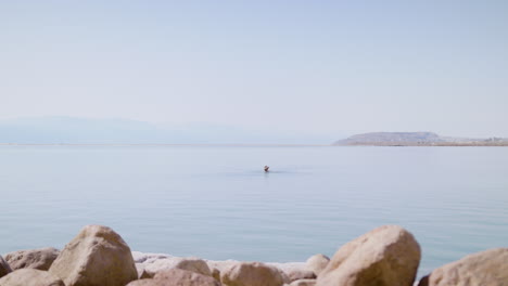 Elderly-tourist-with-a-hat,-enjoys-jumping-and-floating-in-the-Dead-Sea---wide-shot