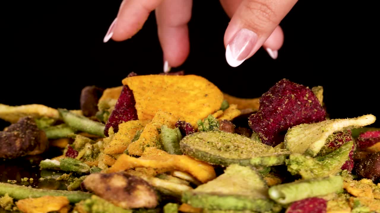 A hand reaches in and picks up assorted dried vegetable chips, including beetroot, potato, mushroom, and beans, under bright studio lighting with a static camera
