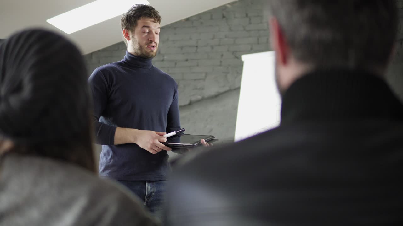 Handsome young businessman pointing at flipchart during presentation in conference room and holding tablet. Presentation speech with flipchart in office. Shot in 4k