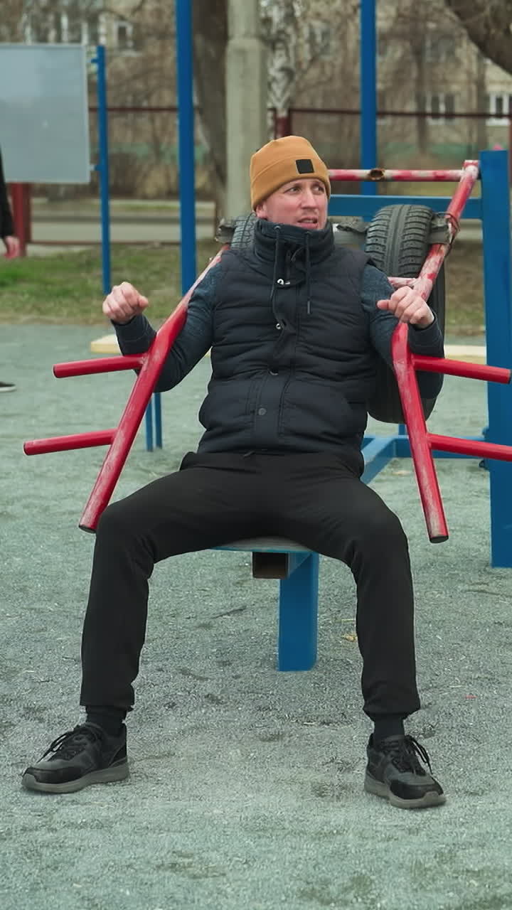 A coach in a brown beanie working out on an outdoor, with two boys in the background playing football, with blue pull up poles around
