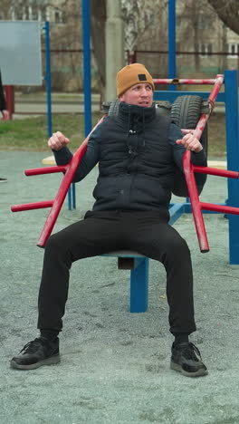 a coach in a brown beanie working out on an outdoor, with two boys in the background playing football, with blue pull up poles around