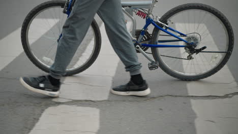 leg view someone in grey pants and sneakers walking alongside a bicycle across a pedestrian crossing, with part of a car approaching, and poles with red markings by the roadside