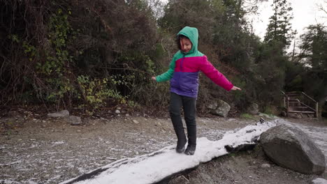 child balancing while walking on a snowy tree trunk wearing colorful jacket in winter forest path