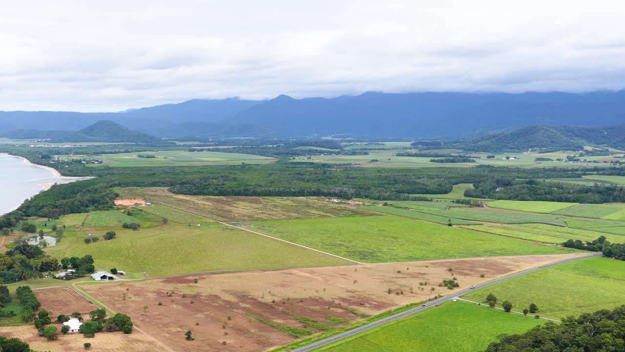Drone glides above Port Douglas coastline, revealing beach, farmland, mountains, and cloudy sky