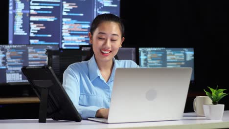 asian female programmer looking at tablet and writing code by a laptop using multiple monitors showing database on terminal window desktops in the office