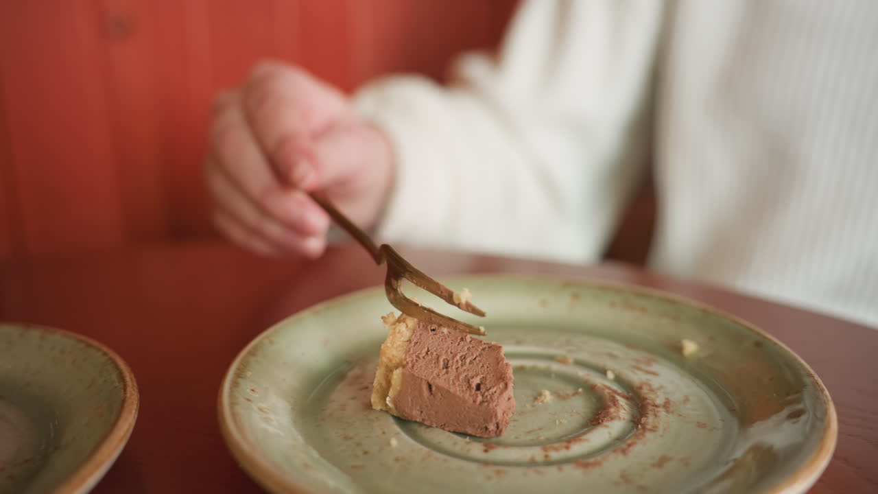 Close up of hand slicing chocolate cake with fork on green ceramic plate beside partially finished latte in transparent glass cup placed on round wooden table in cozy cafe with warm ambient lighting