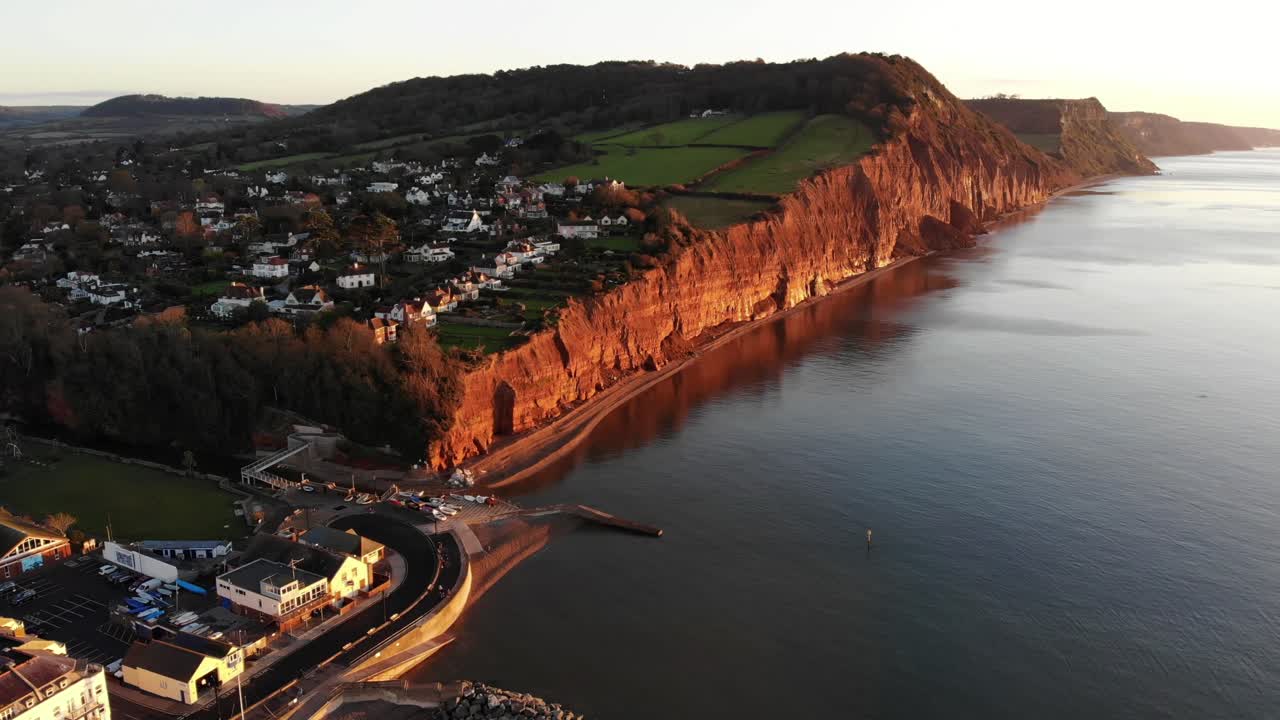Aerial View Of Sidmouth Town Beside Coastline Bathed In Golden Sunrise Light. Dolly Forward Tilt Up