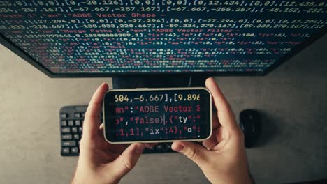 person holding a smartphone displaying code over a computer monitor.