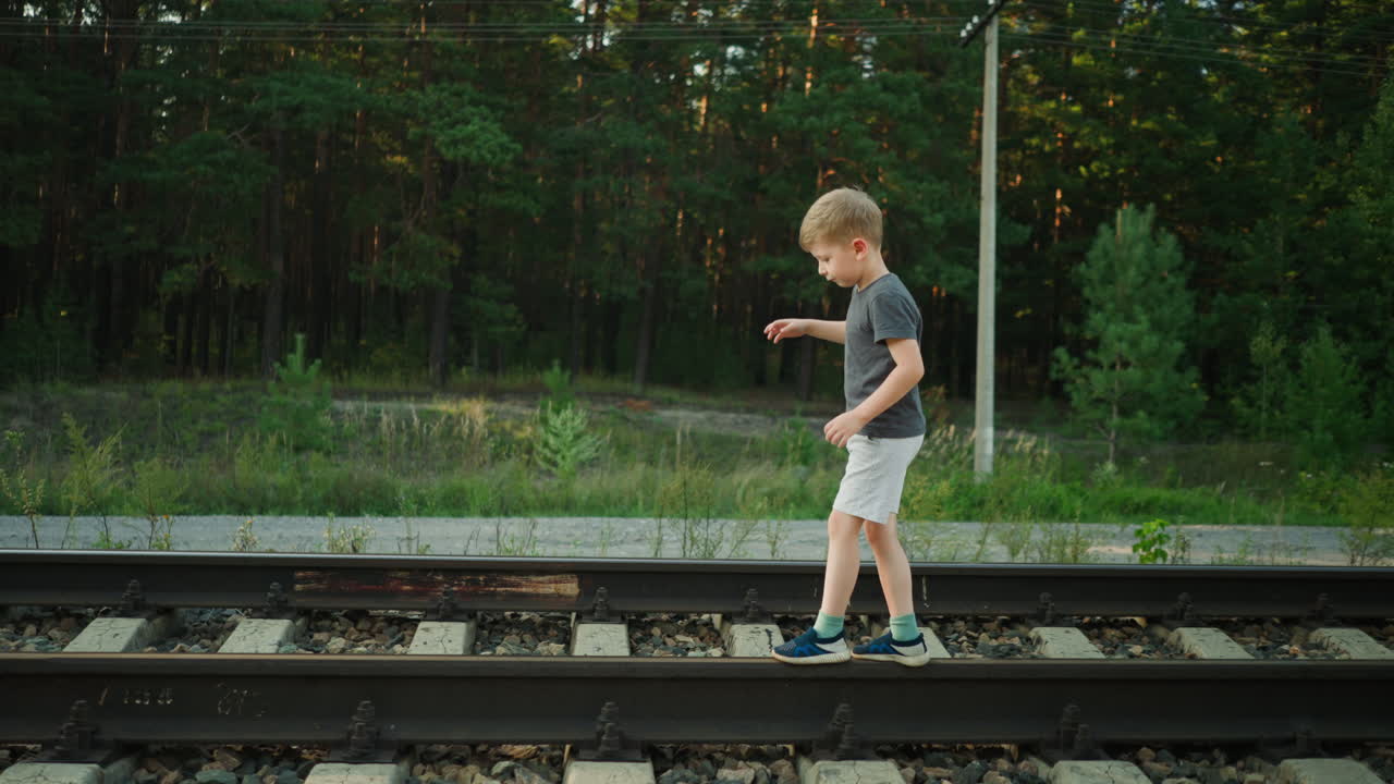 child walking cautiously on rail track surrounded by greenery and dense forest, balancing step by step under soft daylight with power line poles and natural surroundings visible in peaceful setting