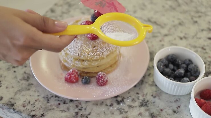 Giovane donna spolvera zucchero su una torre di pancake fatti in casa, decorata con lamponi e mirtilli, cucina casalinga