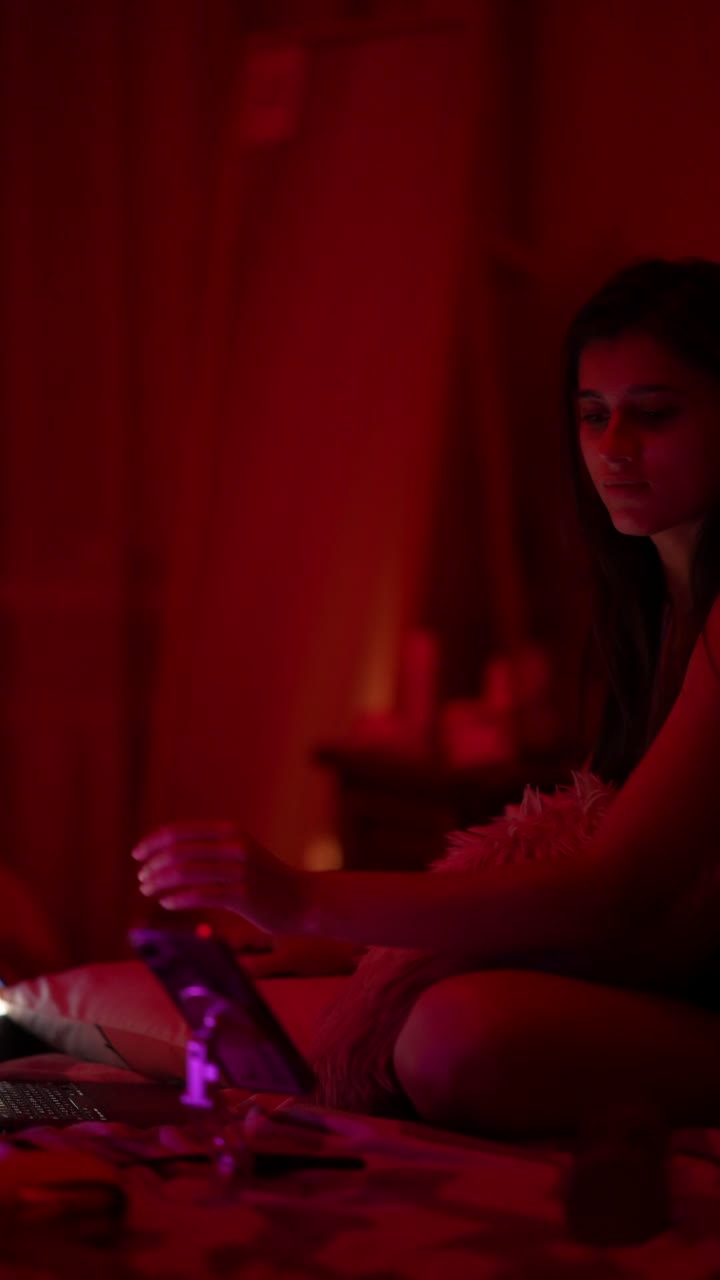 Giovane donna che usa telefono e laptop in una camera da letto illuminata di rosso di notte