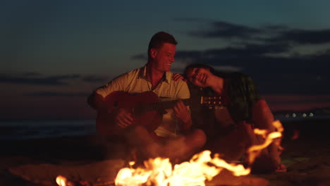 pareja tocando la guitarra junto a la fogata en la playa al atardecer