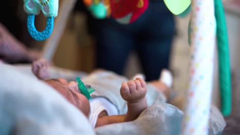 charming newborn baby girl in cradle with soother dummy nipple in mouth moving and staring at toys above her