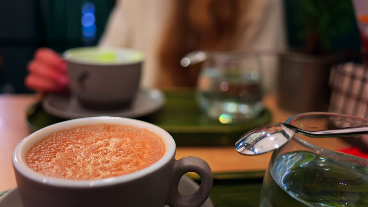 Close up of a cappuccino on a table at a cafe with a blurred view of a woman drinking a matcha latte on the background
