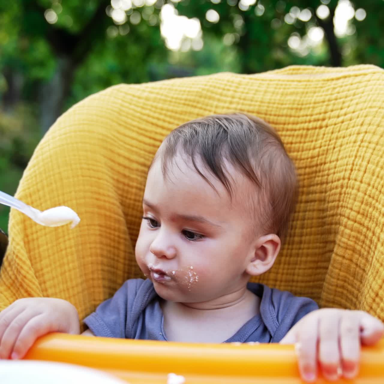 Feeding nice adorable child sitting in the yellow feeding chair. Caring mother feeding her baby from spoon. Close up. Blurred backdrop