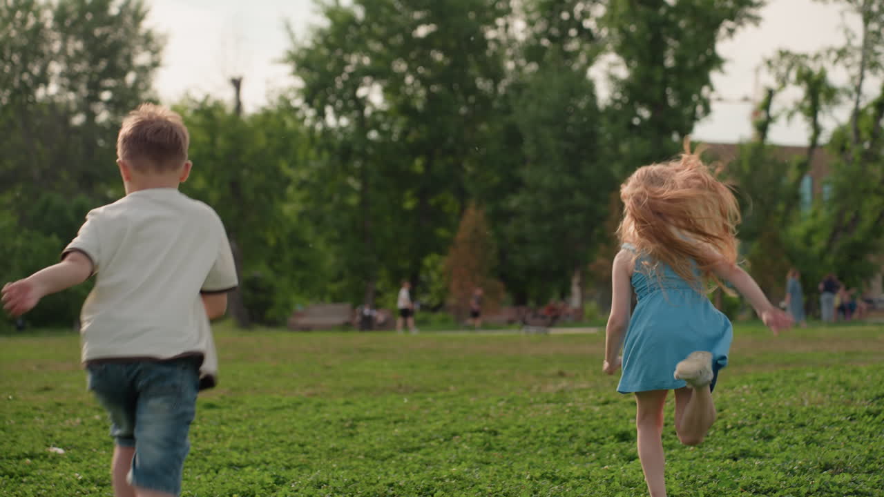 children running across green field in summer park, carefree movement, back view, wind lifting hair, warm light, playful energy, distant adults, nature background capturing joyful outdoor activity