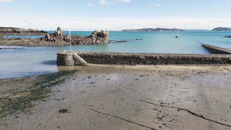 Imágenes-De-Drones-Bajos-Del-Puerto-De-Burdeos,-Guernsey,-Desde-La-Costa-Hacia-La-Pared-Del-Puerto-En-Un-Día-Soleado-Con-Herm-Y-Jethou-Al-Fondo