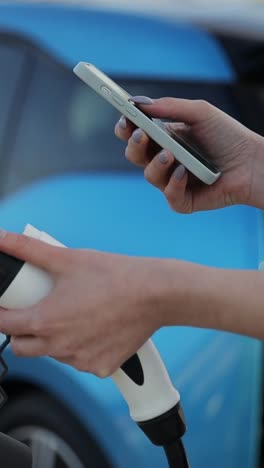 vertical video, woman is standing next to her electric car, charging it while using her cell phone. the vehicles hood is open, showing automotive parts like the tire and wheel