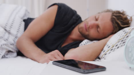 man asleep in bed reaches out to turn off alarm on mobile phone