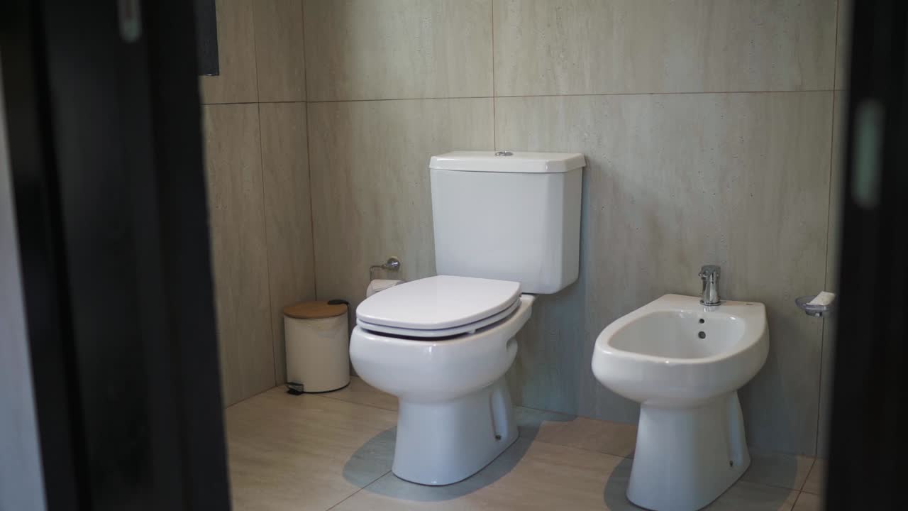 Slow motion camera shot of toilet and bidet in restroom at hotel in Argentina. Clean white ceramic artifacts against cream ceramic wall.