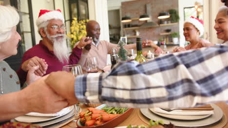 Happy-diverse-senior-friends-holding-hands-to-say-grace-at-christmas-dining-table,-slow-motion