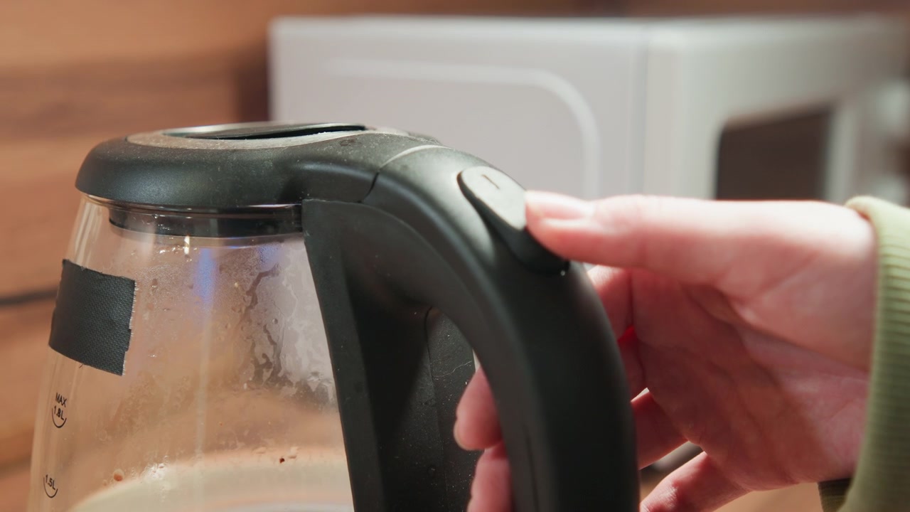 Close three-quarter view shows hand gripping handle and pressing top switch while steam rises inside clear kettle, with condensation, warm light and home kitchen setting, with kettle, hand, glass