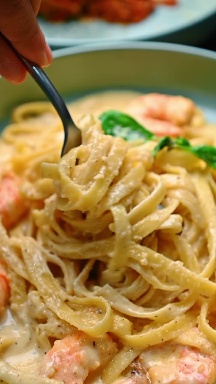 Mano che usa la forchetta per mangiare pasta cremosa con gamberi. Scatto d'azione di una mano che solleva fettuccine con gamberi e basilico da una ciotola azzurra con salsa cremosa.