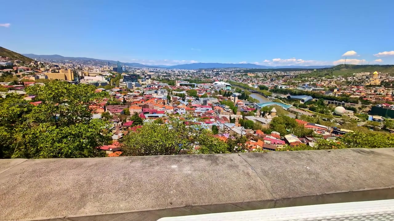 Lavička s výhledem na řeku a panorama Tbilisi pod jasně modrou oblohou.