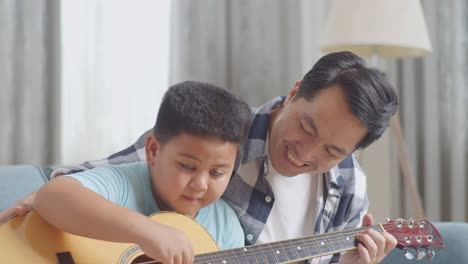 close up of asian father and his son smiling to camera while playing the guitar together on sofa at home