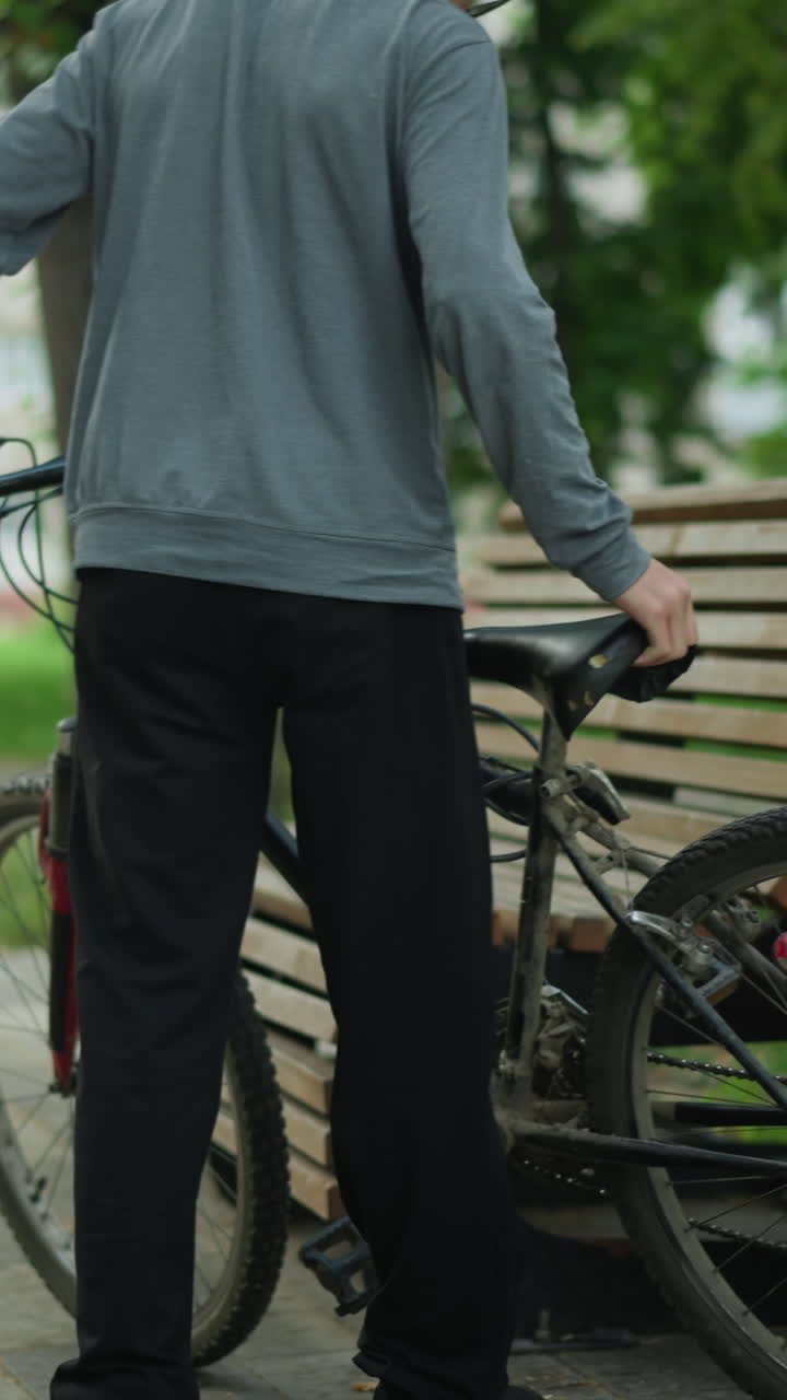 회색  위쪽 안경을 입은 어린 소년은 공원에서 나무 벤치 근처의 자전거를 내려 앉아서 얼굴을 기 전에 벤치에 가까운 자전거을 조정합니다. 나무, 초록색, 도시 배경과 함께