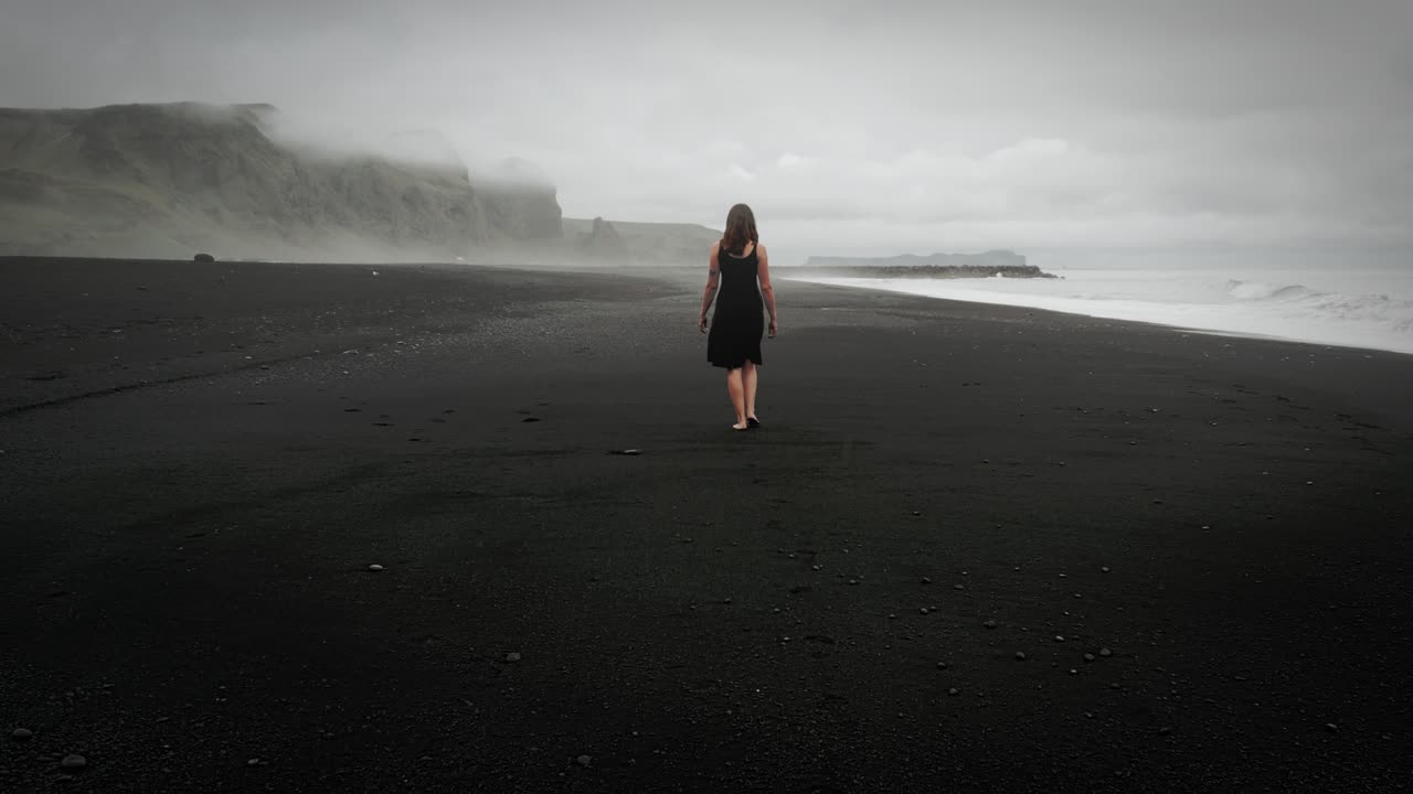 joven hermosa con vestido negro caminando por la playa de arena negra islandesa, paisaje montañoso dramático y brumoso, disparo de seguimiento