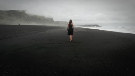 Junge-Schöne-Frau-Im-Schwarzen-Kleid-Spaziert-Am-Schwarzen-Sandstrand-Islands,-Neblige-Dramatische-Berglandschaft,-Kamerafahrt