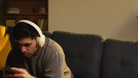 young man spending evening at home sitting on sofa wearing wireless headphones streaming music from mobile phone