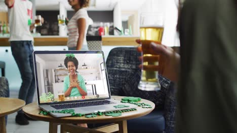 Smiling-african-american-woman-with-beer-wearing-clover-shape-band-on-video-call-on-laptop