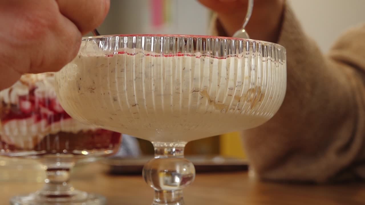 Primer plano de un postre cremoso por capas servido en un bol de cristal decorativo, cubierto con una fina capa de gelatina roja, con otro postre de frutas difuminado al fondo.