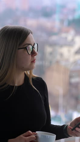 Businesswoman with a phone and a cup in office. Young woman drinking coffee and uses mobile phone on window background with a city view. Vertical video