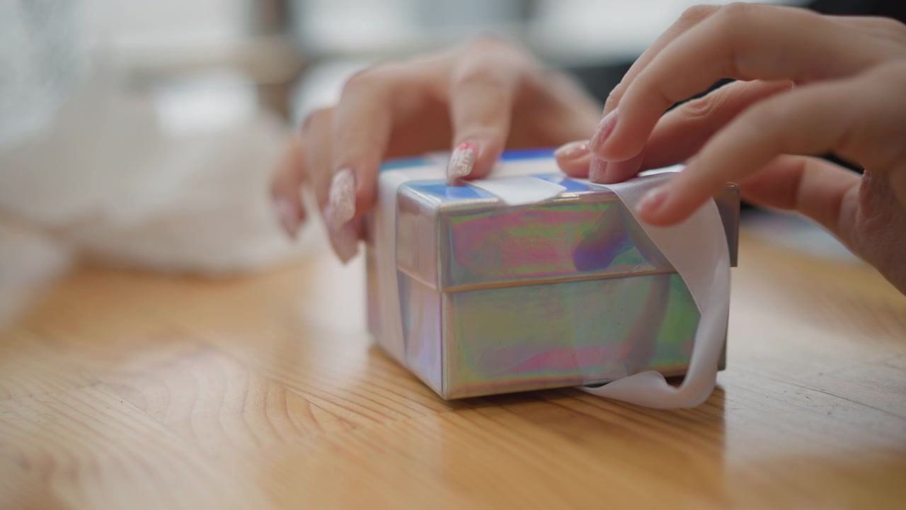 暖かな木製のテーブルに置かれた小さな玉虫色の箱の上で、リボンを引っ張って形を整え、蝶結びにしてギフトラッピングを仕上げる手のクローズアップ。