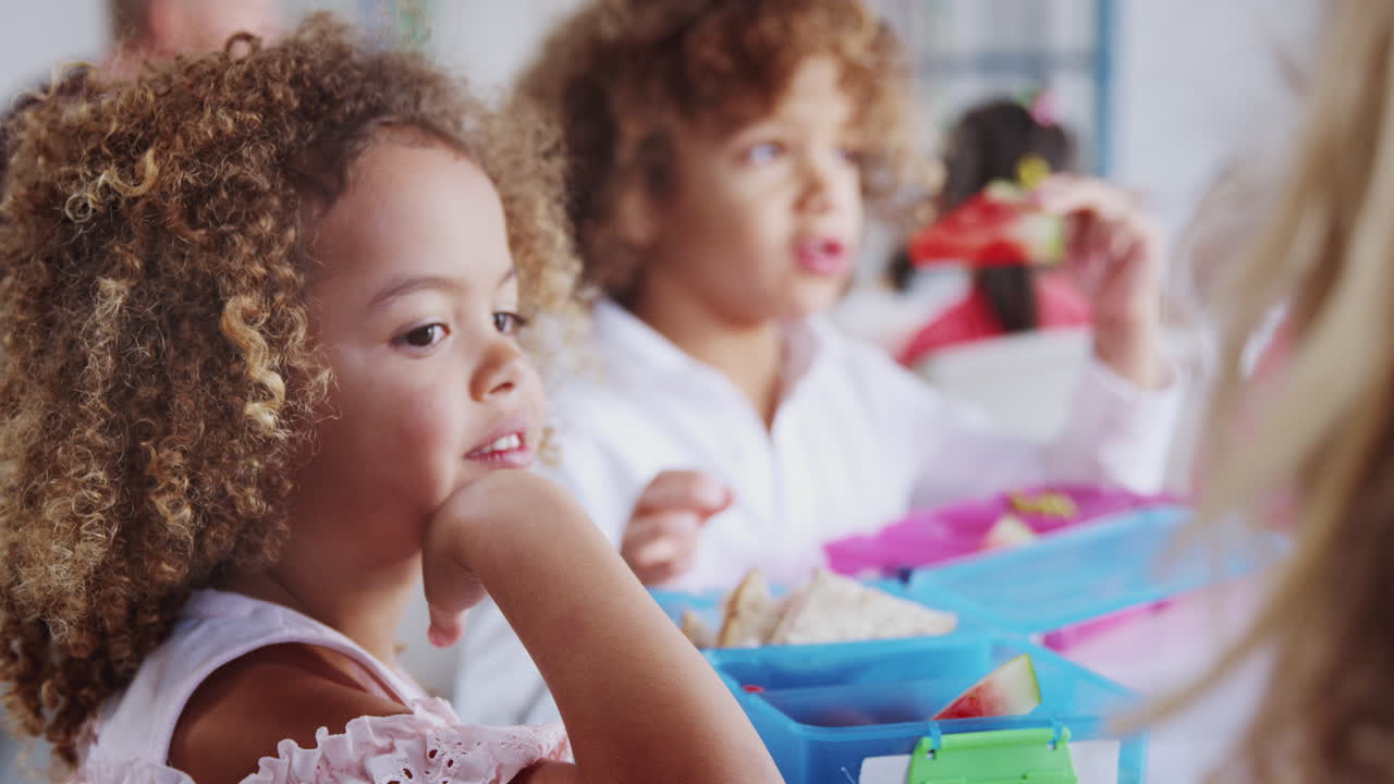 Primo piano di una bambina della scuola dell'infanzia che mangia il suo pranzo al sacco con i compagni di classe, messa a fuoco selettiva.